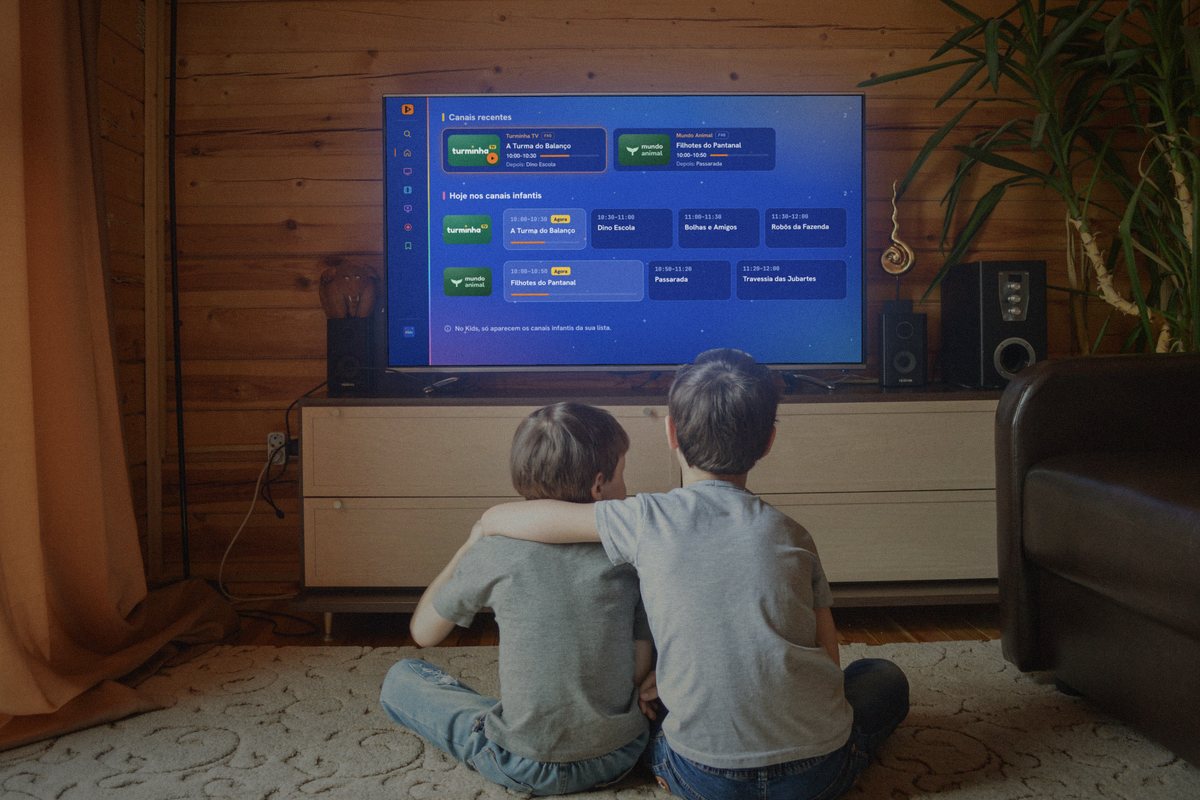Duas crianças de costas no tapete. Na TV, o Kids azul, só com os canais infantis Turminha TV e Mundo Animal.